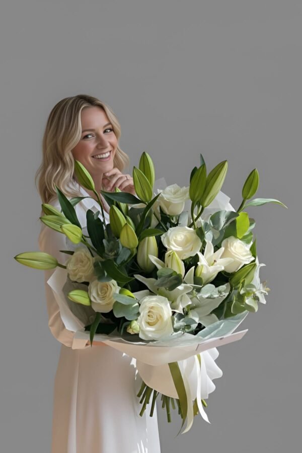 A smiling blonde woman holding a large, luxurious bouquet of white lilies, Avalanche roses, and eucalyptus, hand-wrapped in premium white paper.