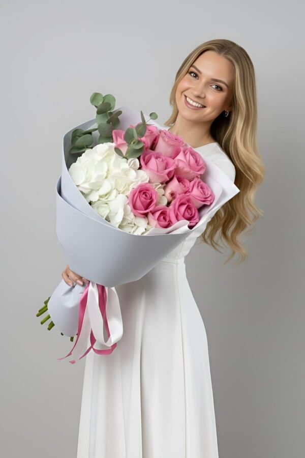 A blonde woman in a white dress holding a luxury bouquet of pink roses, white hydrangeas, and eucalyptus, wrapped in elegant light grey paper with a pink ribbon.